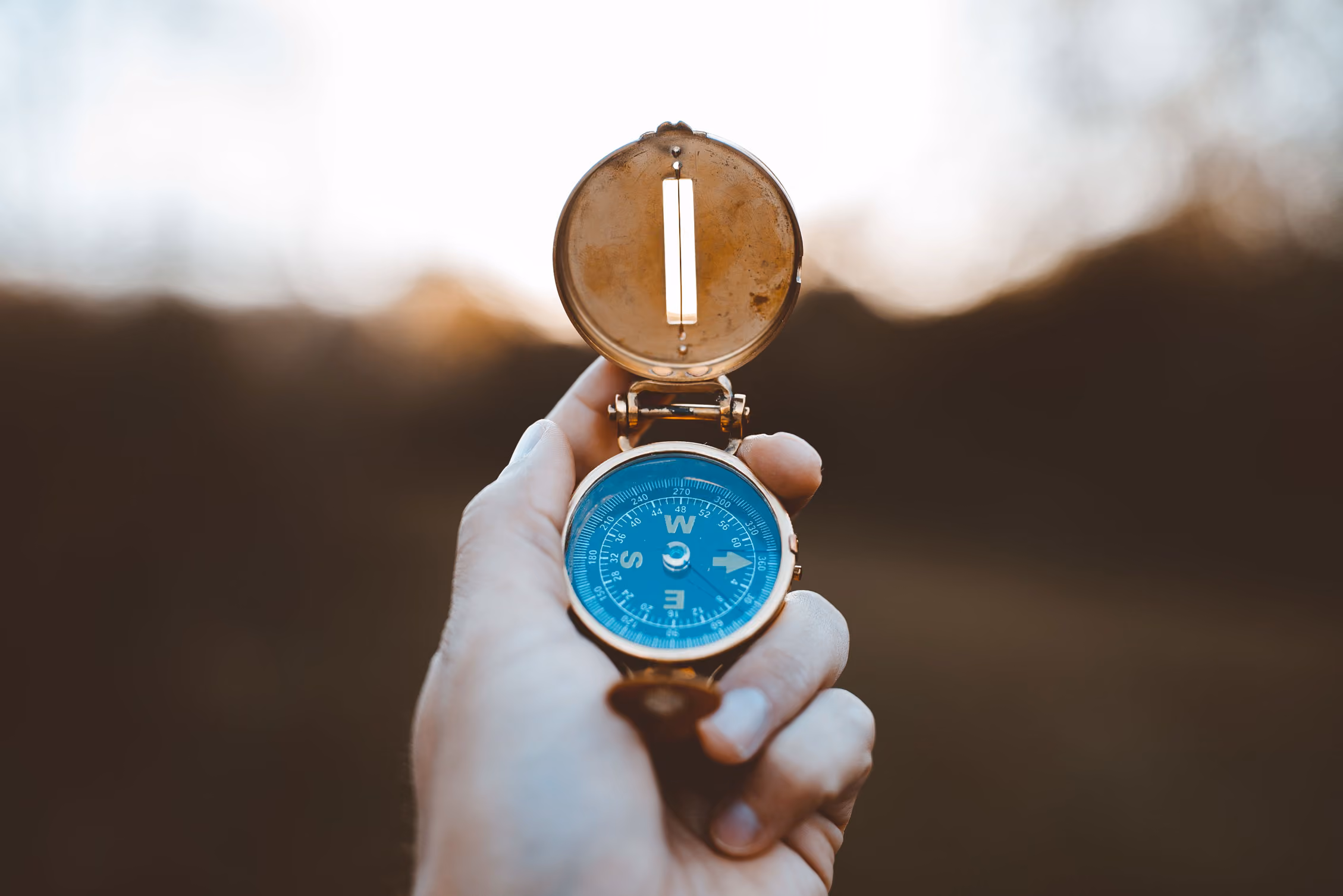 Hand holding an open brass compass with a blue face against a blurred outdoor background.